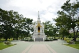 A small, ornate white temple surrounded by green trees and well-maintained gardens. The temple features a golden statue and intricate architectural details. The sky is partially cloudy, and the pathway leading to the temple is tiled in green and beige.