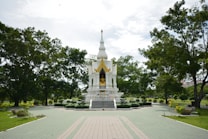 A small, ornate white temple surrounded by green trees and well-maintained gardens. The temple features a golden statue and intricate architectural details. The sky is partially cloudy, and the pathway leading to the temple is tiled in green and beige.