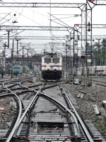 A train approaches on a complex network of railway tracks with overhead wires and numerous poles. The setting is busy with multiple tracks converging and diverging, indicating a railway junction. The background shows more tracks, infrastructure, and some greenery.