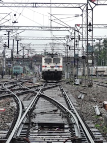 A train approaches on a complex network of railway tracks with overhead wires and numerous poles. The setting is busy with multiple tracks converging and diverging, indicating a railway junction. The background shows more tracks, infrastructure, and some greenery.