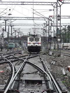 A train approaches on a complex network of railway tracks with overhead wires and numerous poles. The setting is busy with multiple tracks converging and diverging, indicating a railway junction. The background shows more tracks, infrastructure, and some greenery.