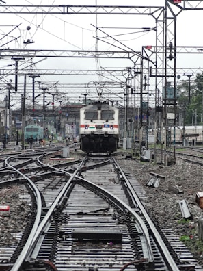 A train approaches on a complex network of railway tracks with overhead wires and numerous poles. The setting is busy with multiple tracks converging and diverging, indicating a railway junction. The background shows more tracks, infrastructure, and some greenery.