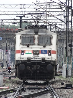 A powerful locomotive is on railway tracks surrounded by complex wiring overhead. The train is stationary, captured from the front view. It features detailed signage and logos, including an Indian flag, and mechanical details like vents and lights. The background shows more tracks, electrical poles, and various signals.