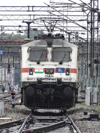 A powerful locomotive is on railway tracks surrounded by complex wiring overhead. The train is stationary, captured from the front view. It features detailed signage and logos, including an Indian flag, and mechanical details like vents and lights. The background shows more tracks, electrical poles, and various signals.