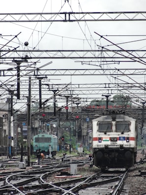 A train station with multiple railway tracks and overhead electrical wires. Two trains are visible, one in the foreground and one farther back, with people walking near the tracks. The scene is urban and industrial, with buildings visible in the background.
