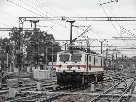 A white and black locomotive is on a busy railway track surrounded by an extensive network of overhead power lines. The background reveals additional train tracks and distant structures, possibly train station facilities, amid a somewhat overcast sky.
