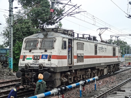 An Indian Railways electric locomotive is positioned on the tracks. The locomotive is white with a red stripe and features the Indian flag. There are overhead electrical wires and a few people are in the vicinity, one of whom is peering out from the driver's cabin.