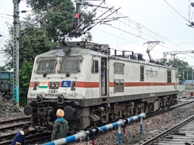 An Indian Railways electric locomotive is positioned on the tracks. The locomotive is white with a red stripe and features the Indian flag. There are overhead electrical wires and a few people are in the vicinity, one of whom is peering out from the driver's cabin.