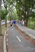 A group of people, including children and adults, walking along a tree-lined path in a park. They appear to be enjoying an outdoor activity, likely on a guided tour or excursion. The path is surrounded by lush greenery with large trees, and there is a small body of water to the right.