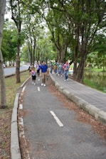 A group of people, including children and adults, walking along a tree-lined path in a park. They appear to be enjoying an outdoor activity, likely on a guided tour or excursion. The path is surrounded by lush greenery with large trees, and there is a small body of water to the right.