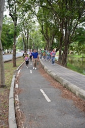 A group of people, including children and adults, walking along a tree-lined path in a park. They appear to be enjoying an outdoor activity, likely on a guided tour or excursion. The path is surrounded by lush greenery with large trees, and there is a small body of water to the right.