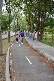 A group of people, including children and adults, walking along a tree-lined path in a park. They appear to be enjoying an outdoor activity, likely on a guided tour or excursion. The path is surrounded by lush greenery with large trees, and there is a small body of water to the right.