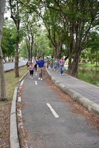 A group of people, including children and adults, walking along a tree-lined path in a park. They appear to be enjoying an outdoor activity, likely on a guided tour or excursion. The path is surrounded by lush greenery with large trees, and there is a small body of water to the right.