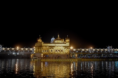An illuminated golden temple is reflected in a serene body of water at night, surrounded by a symmetrical architectural structure with numerous lights.