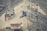 A person wearing a safety harness and a yellow hard hat is sitting on a steep, corrugated metal roof, surrounded by ropes. Mobile antenna towers and other equipment are visible, indicating maintenance or construction work.
