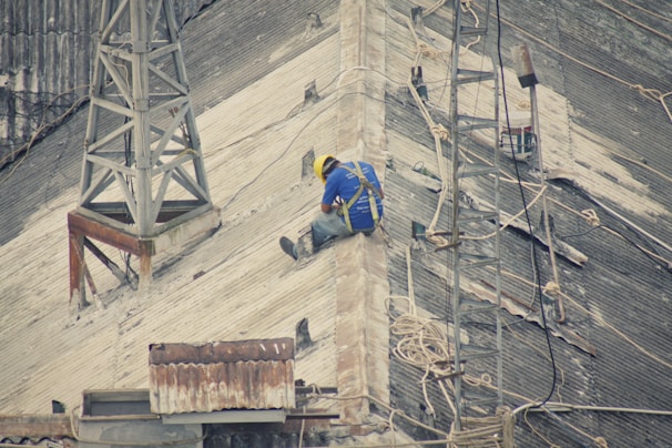 A person wearing a safety harness and a yellow hard hat is sitting on a steep, corrugated metal roof, surrounded by ropes. Mobile antenna towers and other equipment are visible, indicating maintenance or construction work.