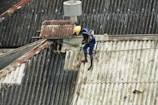 A person is working on a corrugated metal roof, wearing a harness for safety. They appear to be engaged in some kind of maintenance or repair work, holding equipment or tools. The roof is weathered and shows patches of rust and wear.