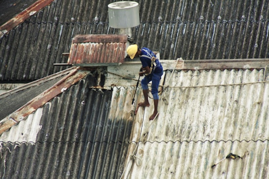 A person is working on a corrugated metal roof, wearing a harness for safety. They appear to be engaged in some kind of maintenance or repair work, holding equipment or tools. The roof is weathered and shows patches of rust and wear.
