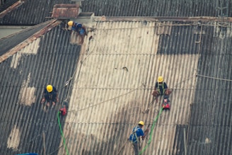 Workers wearing safety harnesses and yellow helmets are performing maintenance on a large corrugated metal roof. They are using equipment attached to ropes secured on the roof.