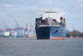 A large cargo ship filled with numerous shipping containers is floating on the water in a harbor. In the background, there are multiple cranes, other ships, and industrial structures lining the port with a slightly overcast sky.