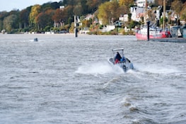 A small motorboat speeds across a river, creating a trail of white water behind it. The river is wide, with choppy waves, and lined by a lush landscape of trees in autumn colors. Modern houses and buildings are visible along the bank. A red and white boat is docked near the side, while a smaller vessel is seen further in the background. Several people are present on a distant path parallel to the river.