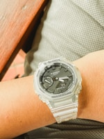 A person's wrist is adorned with a Casio G-Shock watch featuring a transparent strap and a black face. The watch has markings for protection, mode, adjust, and other functions. The background includes a gray surface, likely fabric, and a wooden edge of a table.