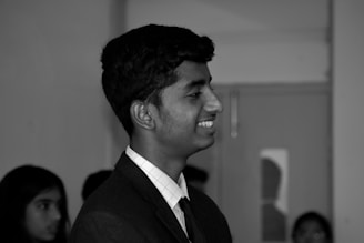 A young person is shown in profile view, smiling. The individual is wearing a formal suit with a tie. Other people are blurred in the background, creating a focus on the central figure.