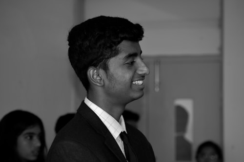 A young person is shown in profile view, smiling. The individual is wearing a formal suit with a tie. Other people are blurred in the background, creating a focus on the central figure.