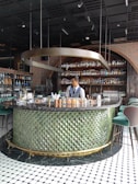 A bartender stands behind a circular bar counter with a textured green exterior and a black marble top. The background features shelves lined with various bottles, likely alcoholic beverages. Overhead, a unique metallic spiral fixture hangs from the ceiling, contributing to the stylish and modern ambiance. The floor is tiled with a black and white geometric pattern.