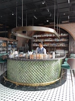 A bartender stands behind a circular bar counter with a textured green exterior and a black marble top. The background features shelves lined with various bottles, likely alcoholic beverages. Overhead, a unique metallic spiral fixture hangs from the ceiling, contributing to the stylish and modern ambiance. The floor is tiled with a black and white geometric pattern.