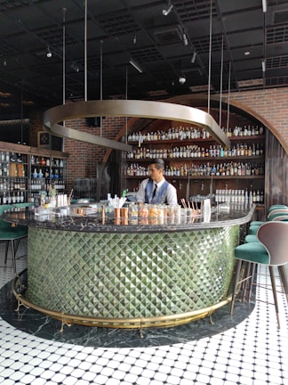 A bartender stands behind a circular bar counter with a textured green exterior and a black marble top. The background features shelves lined with various bottles, likely alcoholic beverages. Overhead, a unique metallic spiral fixture hangs from the ceiling, contributing to the stylish and modern ambiance. The floor is tiled with a black and white geometric pattern.