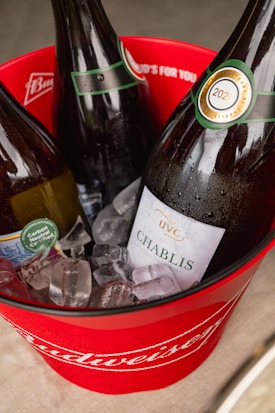 Three wine bottles chilled in ice are placed inside a branded red Budweiser bucket. The bottles have labels with green accents, and the label on one of them reads 'Chablis' with a vintage year of 2021. The setting appears casual, possibly part of a social event or gathering.