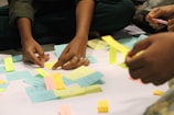 Several hands are reaching towards a collection of colorful sticky notes arranged on a flat, white surface. The sticky notes are in pastel shades of yellow, blue, pink, and green, with handwritten text on each. The activity suggests brainstorming or planning, possibly in a team setting, with individuals collaborating and engaging with the notes.
