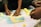 Several hands are reaching towards a collection of colorful sticky notes arranged on a flat, white surface. The sticky notes are in pastel shades of yellow, blue, pink, and green, with handwritten text on each. The activity suggests brainstorming or planning, possibly in a team setting, with individuals collaborating and engaging with the notes.