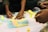 Several hands are reaching towards a collection of colorful sticky notes arranged on a flat, white surface. The sticky notes are in pastel shades of yellow, blue, pink, and green, with handwritten text on each. The activity suggests brainstorming or planning, possibly in a team setting, with individuals collaborating and engaging with the notes.