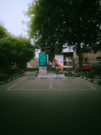 An outdoor parking area with designated spaces marked on the ground. Two large advertisement banners are visible, partially behind a metal fence with trees and foliage in the background. Part of the banners include a digital display and some branding elements. Decorative plants in pots line the sides, with no vehicles or people present.