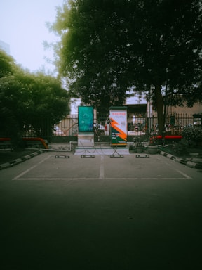 An outdoor parking area with designated spaces marked on the ground. Two large advertisement banners are visible, partially behind a metal fence with trees and foliage in the background. Part of the banners include a digital display and some branding elements. Decorative plants in pots line the sides, with no vehicles or people present.