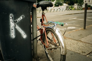 A bicycle with a red frame is chained to a pole on a city street. The bike has a black seat labeled 'Comfort' and a silver fender. A trash bin with a large painted letter 'K' is visible next to it. The background shows parked bicycles and a street with a sidewalk.