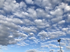 A sky filled with fluffy clouds scattered across a bright blue background. In the lower right corner, a tall pole with a camera or sensor attached is visible.