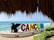Large colorful letters spelling 'CANCUN' stand prominently near a beach. A black silhouette of a winged creature sits beside the letters. Behind them, clear blue ocean waters and a partly cloudy sky create a tranquil backdrop, while lush greenery flanks the scene. A thatched roof at the top of the image provides a shaded frame for the beach view.