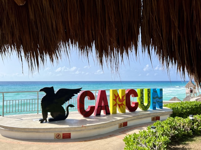 Large colorful letters spelling 'CANCUN' stand prominently near a beach. A black silhouette of a winged creature sits beside the letters. Behind them, clear blue ocean waters and a partly cloudy sky create a tranquil backdrop, while lush greenery flanks the scene. A thatched roof at the top of the image provides a shaded frame for the beach view.