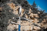 A small wind turbine stands in the foreground, set against a rocky hillside with sparse vegetation and trees. Another turbine can be seen in the distance. The scene suggests renewable energy installations in a natural environment.