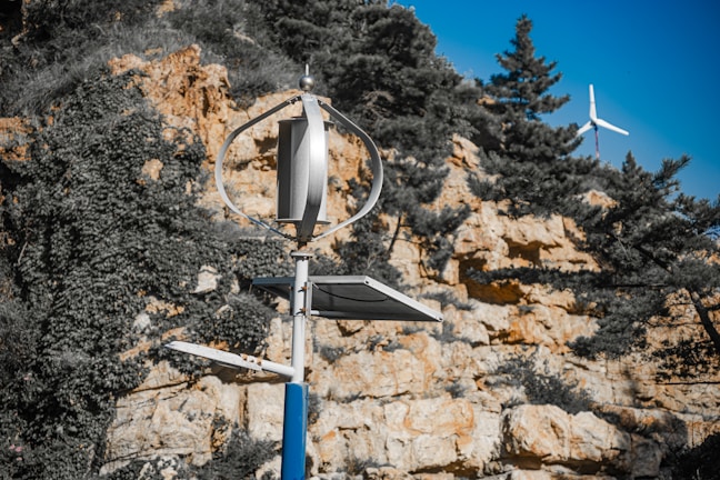A small wind turbine stands in the foreground, set against a rocky hillside with sparse vegetation and trees. Another turbine can be seen in the distance. The scene suggests renewable energy installations in a natural environment.