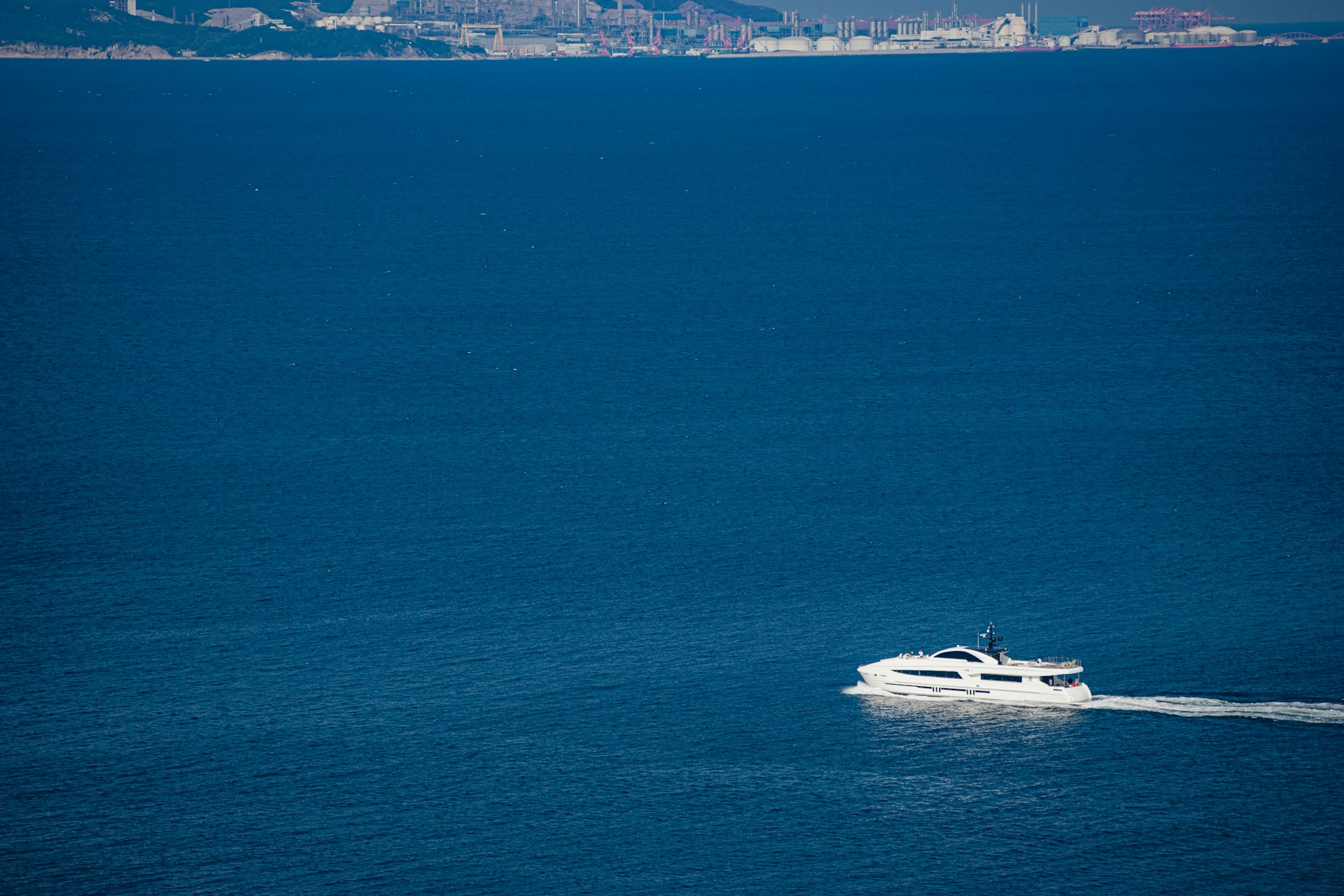 A white yacht is cruising through expansive blue waters with a distant coastal industrial area visible on the horizon.