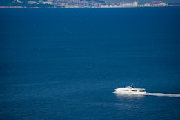 A white yacht is cruising through expansive blue waters with a distant coastal industrial area visible on the horizon.