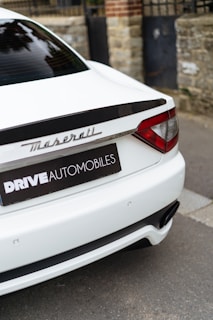 The rear of a luxury white car with the Maserati logo, parked next to a stone wall and metal gate. The branding 'DRIVE AUTOMOBILES' is visible on a black license plate frame.