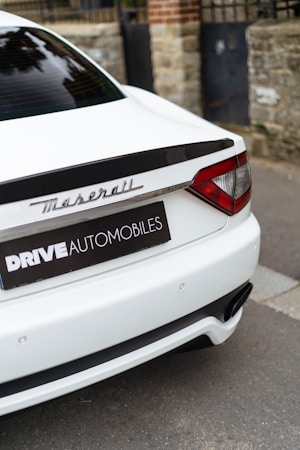 The rear of a luxury white car with the Maserati logo, parked next to a stone wall and metal gate. The branding 'DRIVE AUTOMOBILES' is visible on a black license plate frame.