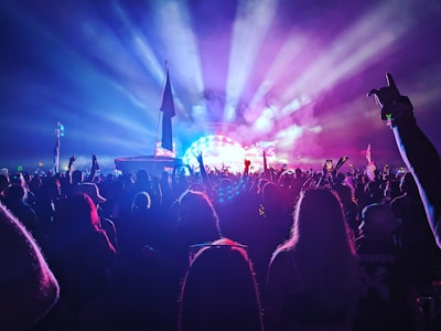 A large crowd of people enjoying a concert or music festival at night. Colorful stage lights beam outwards from the stage, creating a vibrant and energetic atmosphere. Some attendees are holding flags or banners and are silhouetted against the lights.