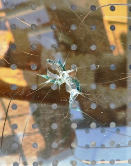 A shattered glass surface with a circular pattern of black dots. The cracks radiate outward from the central impact point, where the glass has splintered significantly. Reflections of a building and sky can be seen in the background, suggesting the glass is part of a window.