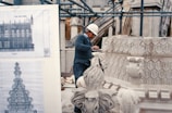 A worker wearing a hard hat is carving intricate designs on a large stone architectural piece. In the background, architectural blueprints and scaffolding can be seen, indicating a construction or restoration site.